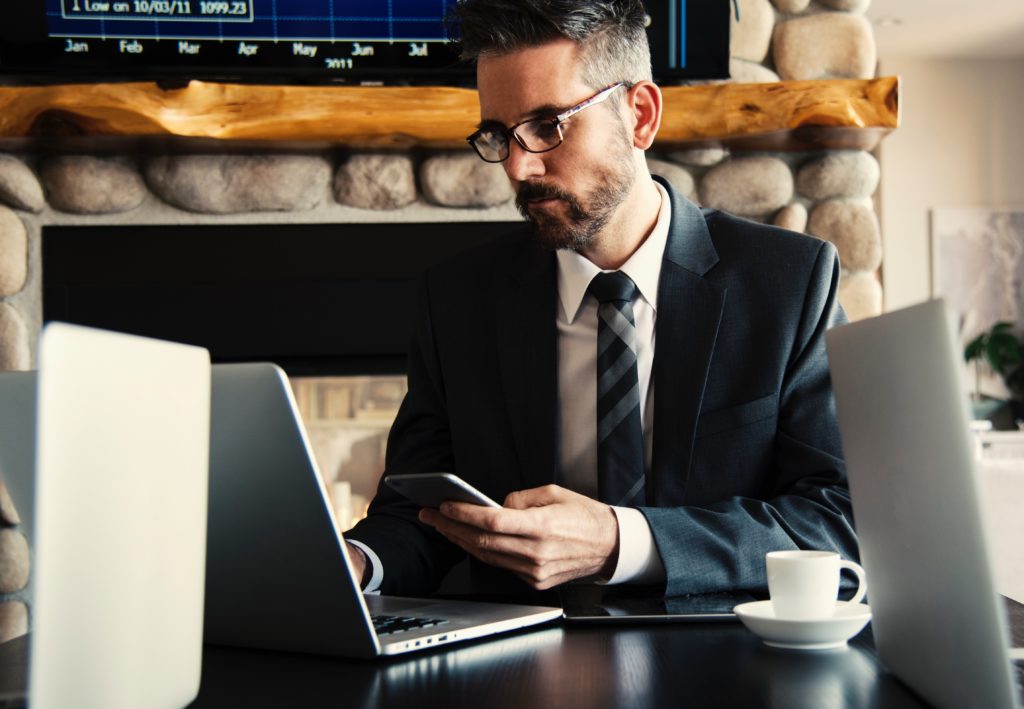 A man updating a CRM on a laptop, whilst holding his phone in a coffee shop.